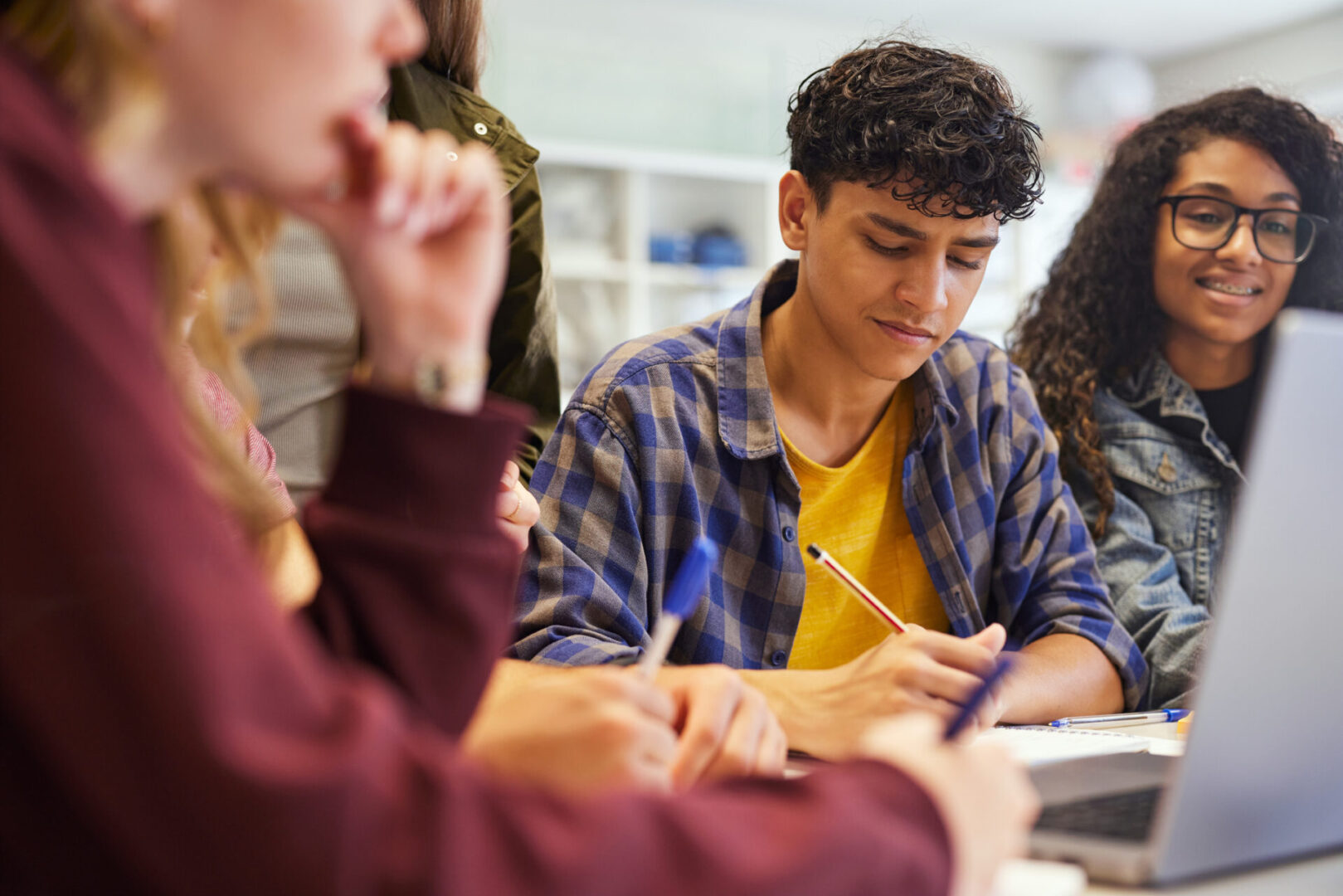 Stressed guy with student friends writing notes during study group learning session. Closeup of multiethnic teens working together during an academic discussion. Classmates concentrating on task while others collaborate beside laptop.